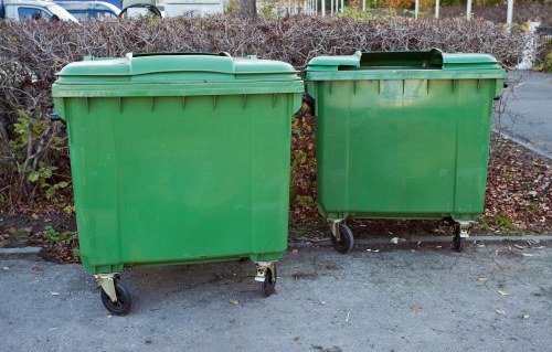 Two large green wheeled rubbish bins positioned side by side on a paved area, possibly an industrial or residential driveway in Tower Hamlets, with their lids closed. The bins are made of durable plastic with textured surfaces and are designed for waste collection and disposal. Behind the bins, there is a row of shrubbery and a metal fence, suggesting an outdoor setting within an urban environment. The pavement surface is slightly worn with a few scattered leaves, typical of an area used for waste management activities. The overall scene reflects a typical site for rubbish removal services provided by Office Clearance Tower Hamlets, emphasizing the importance of proper waste containment and collection in the local community.