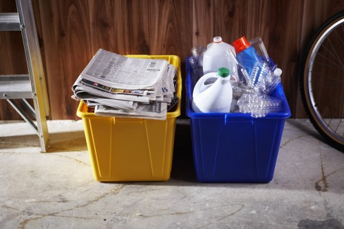 Two large plastic recycling bins are positioned side by side on a concrete floor, one yellow and the other blue. The yellow bin is filled with a stack of newspapers and paper documents, while the blue bin contains several empty or partially filled transparent plastic bottles, as well as a white plastic jug and a clear plastic container. The bins are situated indoors against a wooden-paneled wall, with part of a metal shelving unit visible on the left. To the right, a portion of a bicycle wheel is partially shown, suggesting the location may be a garage or storage area within a property in Tower Hamlets. The environment appears clean and organized, and the presence of the bins indicates ongoing waste or recycling management, consistent with rubbish removal services offered by Office Clearance Tower Hamlets to local residents or businesses in the area, including postal code E1.