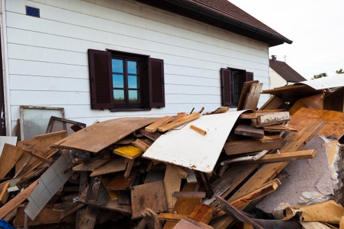 A large pile of assorted construction debris and discarded wooden planks situated beside a white residential building with dark brown window shutters. The debris includes broken wooden boards, particleboard, and pieces of insulation, some leaning and stacked irregularly on the pavement. The background shows the exterior wall of the house, with three windows visible, each with matching dark shutters. The scene depicts a typical outdoor rubbish accumulation possibly in need of removal services by a company like Office Clearance Tower Hamlets, servicing local properties in London’s East End. Natural daylight illuminates the clutter, emphasizing the rough textures and earthy tones of the materials, set against the clean, white finish of the house that contrasts sharply with the chaotic heap of refuse near a paved walkway or driveway area.