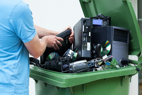 A person wearing a light blue T-shirt is holding a black electronic device near the open lid of a large green wheelie bin. Inside the bin, there is a variety of electronic waste including a desktop computer tower with a black case and a blue-lit internal component, multiple CD-ROM or DVD discs, a digital camera, a pair of black headphones, and several circuit boards and cables. The background shows an outdoor setting with a paved surface, typical of a residential or commercial area in Tower Hamlets, suggesting a rubbish removal or electronic waste disposal service provided by Office Clearance Tower Hamlets. The image emphasizes the collection and disposal of electronic waste, aligned with professional rubbish removal for technical and electronic items in the local area.