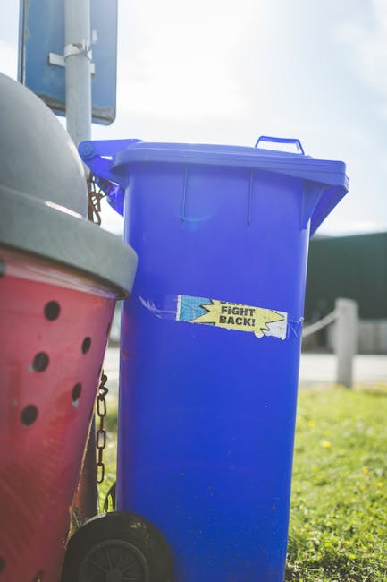 A tall, bright blue wheeled rubbish bin with a closed lid, positioned outdoors on a grassy area, partially in front of a red container with a black lid that has circular ventilation holes. The blue bi