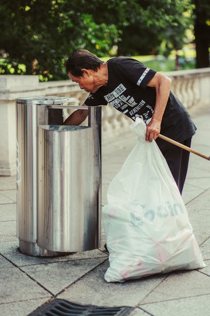 A tall, bright blue wheeled rubbish bin with a closed lid, positioned outdoors on a grassy area, partially in front of a red container with a black lid that has circular ventilation holes. The blue bi